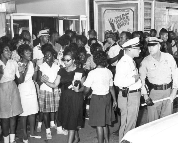 Patricia Stephens Due (in black dress with dark glasses) and John Due (visible above the policeman's cap) are among the protesters outside a segregated movie theater in Tallahassee. Photo from the State Archives of Florida: Florida Memory, http://floridamemory.com/items/show/34010